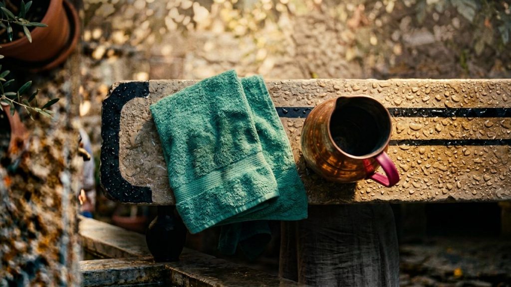 A still-life of a damp folded towel and a copper jug beside a basin with water droplets on the rim, illustrating the Dua After Wudu.