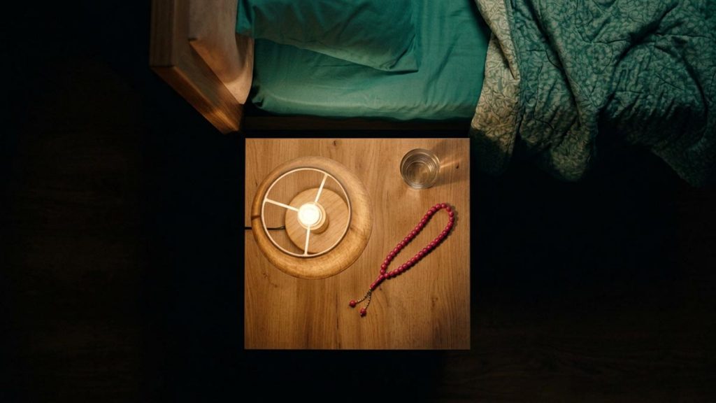 A still-life of a bedside lamp glowing low beside rumpled bedding and prayer beads, illustrating the Dua Against Nightmares.
