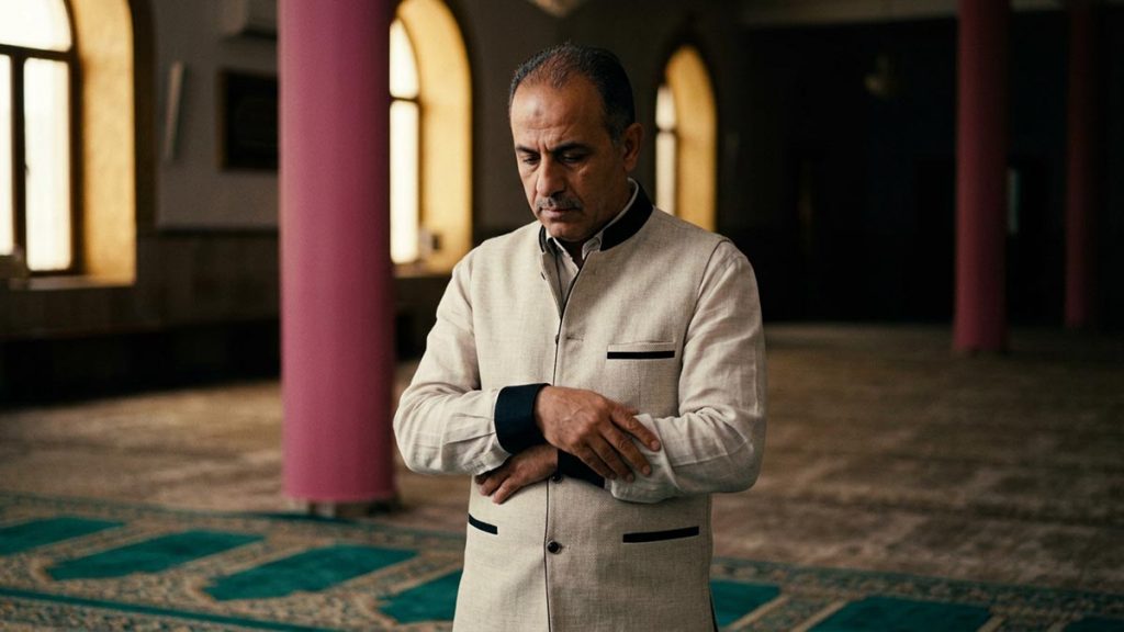 A Muslim man standing at the head of a prayer mat in a quiet hall, an opening scene for the Dua al-Istiftah.