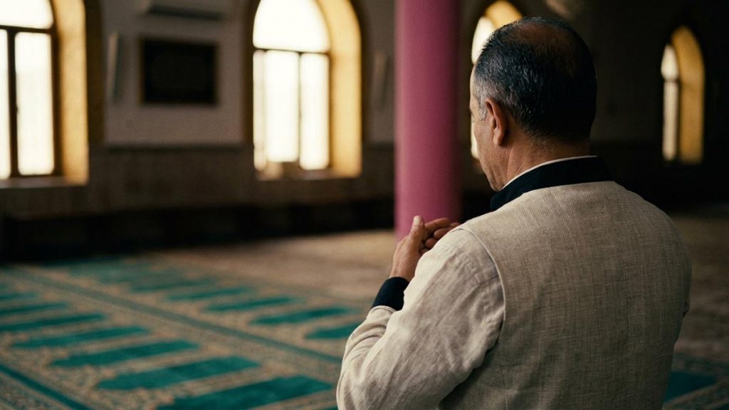 A Muslim man seen from behind standing at the head of a prayer mat in a quiet hall, reflecting the Dua al-Istiftah.