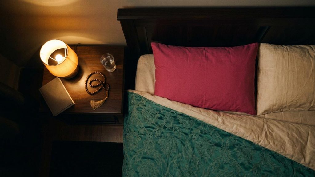 A still-life of turned-down bedding, prayer beads and a small lamp on a bedside table at night, illustrating the Dua Before Sleeping.
