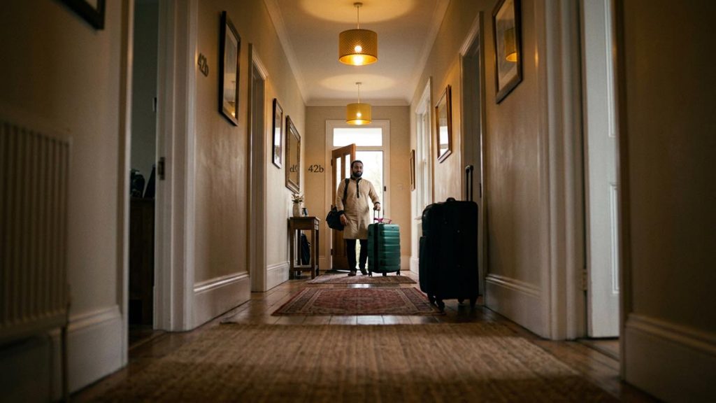 A Muslim man in a hallway with a packed suitcase by the door, an opening scene for the Dua Before Travelling.