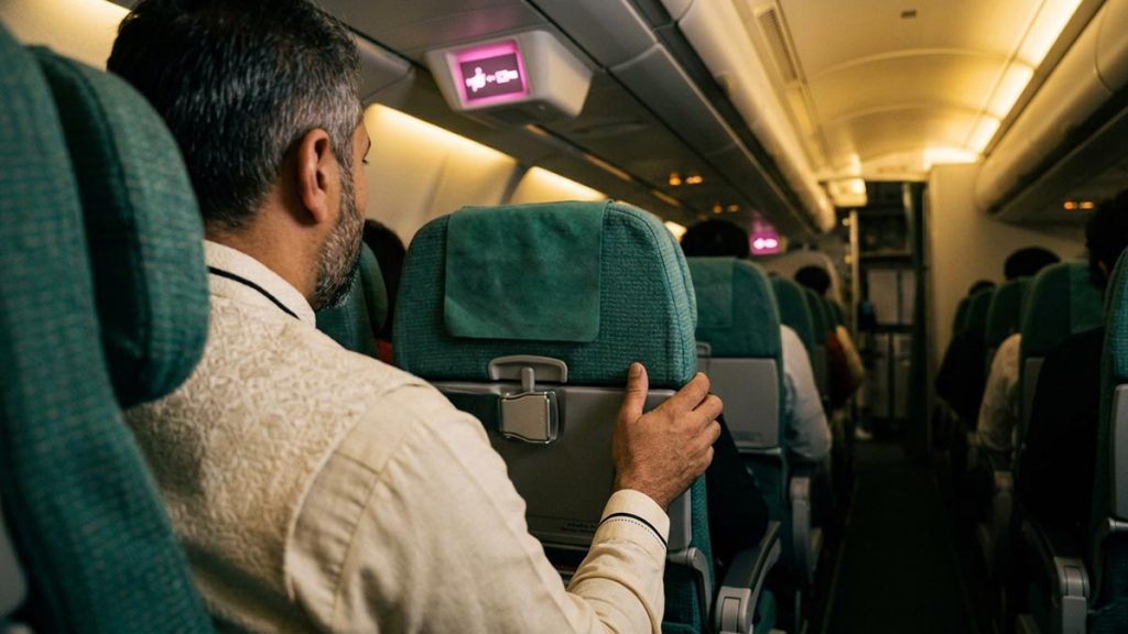 A Muslim man seen from behind in an aircraft cabin seat with the seatbelt sign lit, reflecting the Dua During Turbulence.