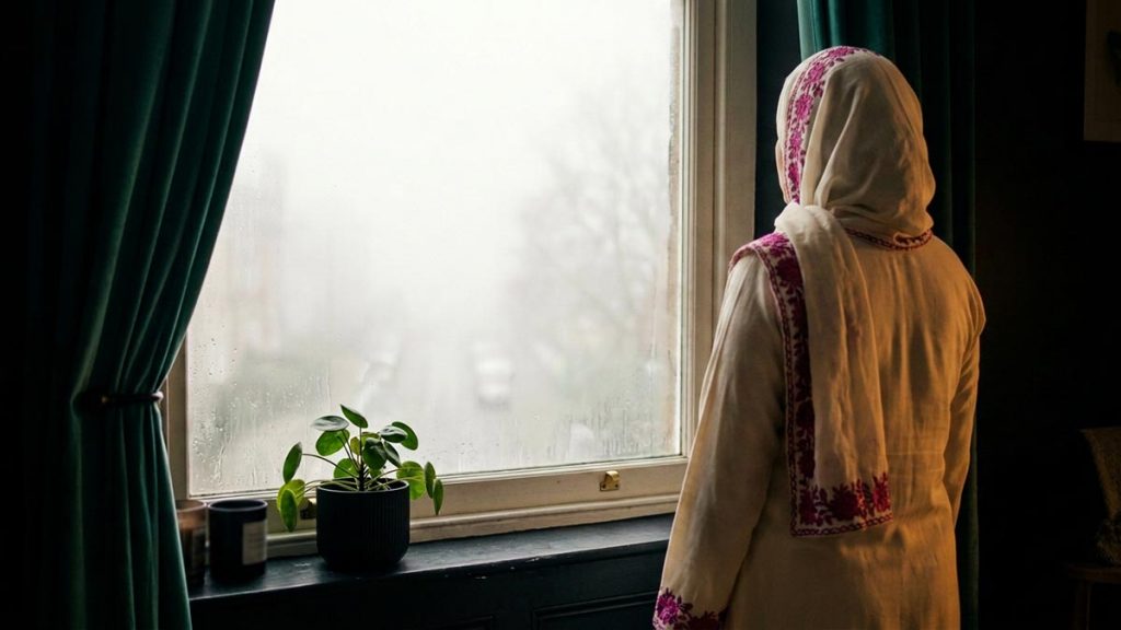 A Muslim woman seen from behind at a window looking out on a fog-covered morning, reflecting the Dua During Uncertainty.
