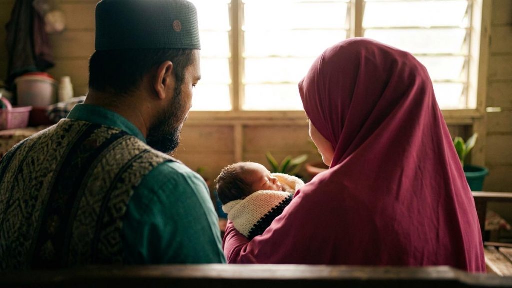 A Muslim family seen from behind in a bright room with a newborn wrapped in a soft blanket, reflecting the Dua for a Newborn Baby.