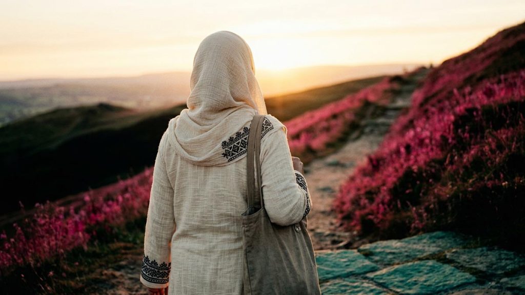 A Muslim woman seen from behind on a hillside path climbing towards a bright horizon, reflecting the Dua for Higher Ranks in Jannah.