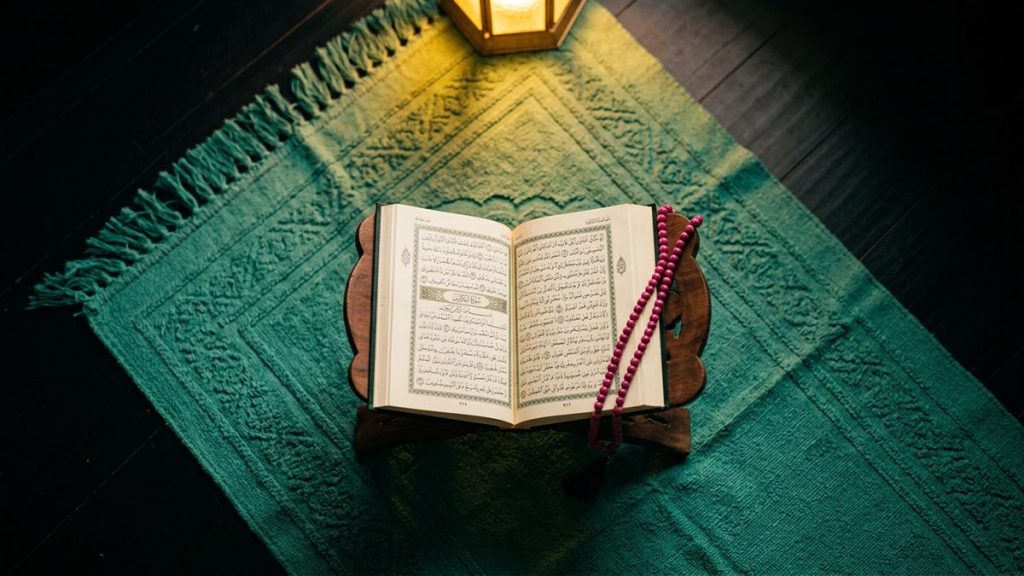 A still-life of a prayer mat with an open Quran and prayer beads laid upon it, illustrating the Dua for Protection from Shaytan.