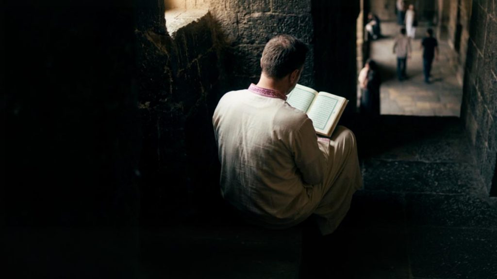 A Muslim man seen from behind in a sheltered stone alcove with the Quran open, reflecting the Dua of the People of the Cave.