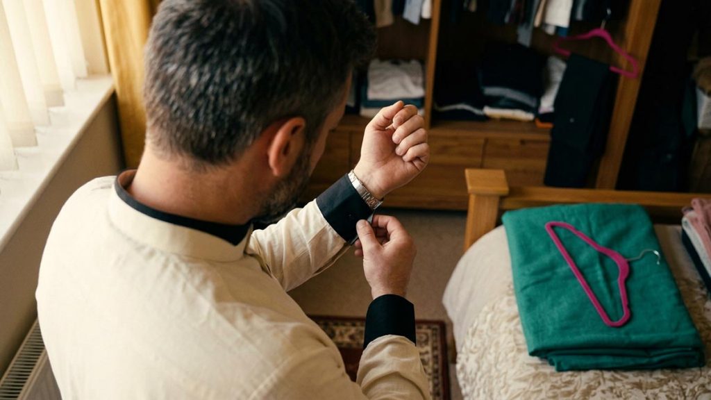 A Muslim man seen from behind in a bedroom beside a wardrobe of neatly hung clothes, reflecting the Dua Upon Wearing New Clothes.