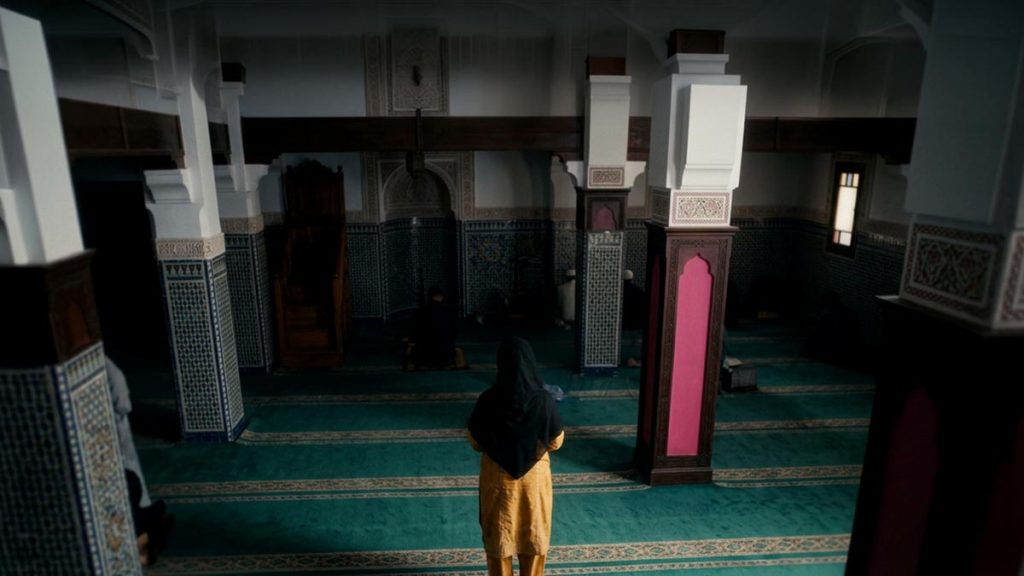 A Muslim woman seen from behind standing on a prayer mat with palms open before the chest, reflecting the Qunoot Dua.